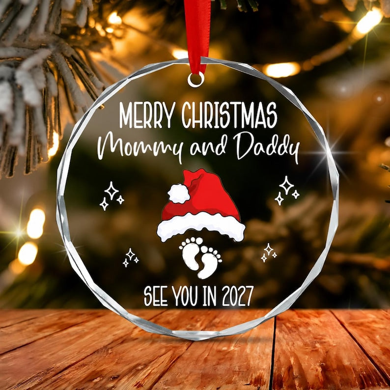 A circular clear glass Christmas ornament with a red ribbon hanger. The text reads MERRY CHRISTMAS Mommy and Daddy, and SEE YOU IN 2027. A red and white Santa hat sits above a pair of white baby footprints with four-pointed stars.