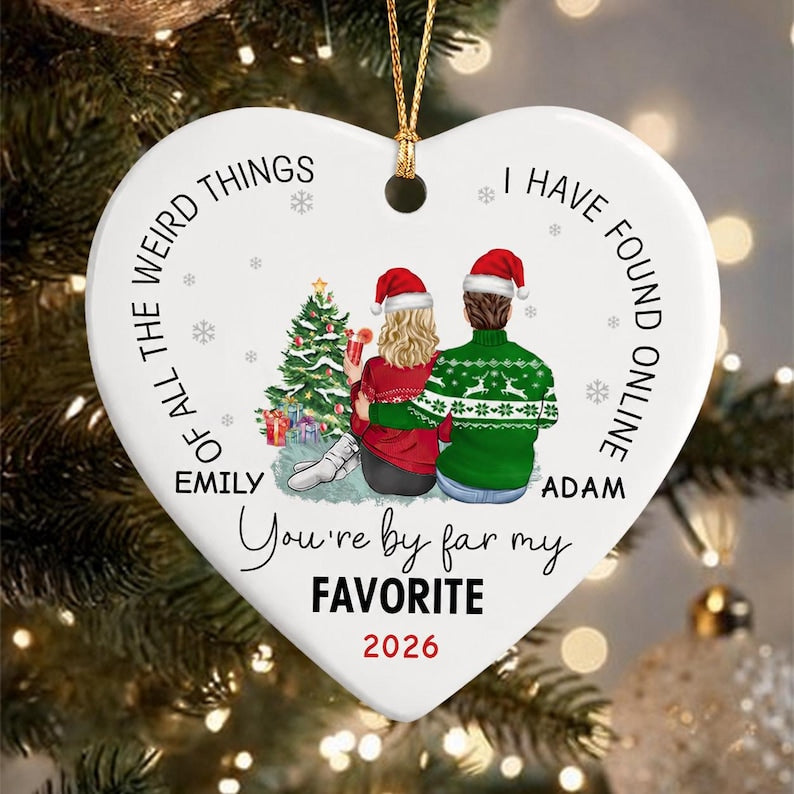 A white heart-shaped ceramic ornament featuring a Christmas tree and two figures in winter attire. Text reads: OF ALL THE WEIRD THINGS I HAVE FOUND ONLINE EMILY ADAM You're by far my FAVORITE 2026. It hangs from a gold cord.
