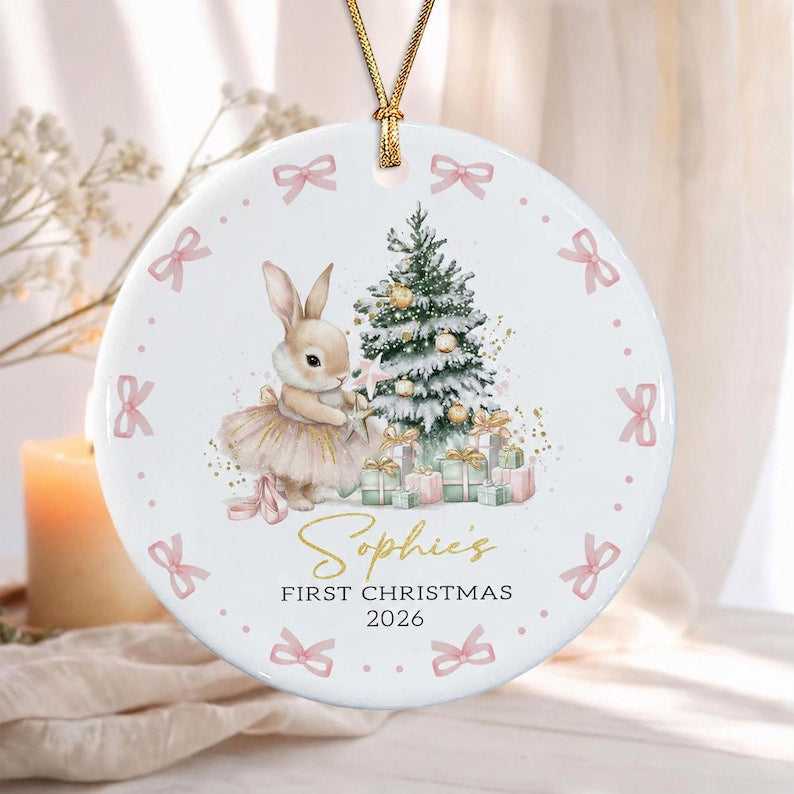 A round white ceramic ornament featuring a gold string hanger. The design includes a rabbit in a tutu, a Christmas tree, and gifts. The text reads Sophie's FIRST CHRISTMAS 2026. The border is decorated with small pink bows.