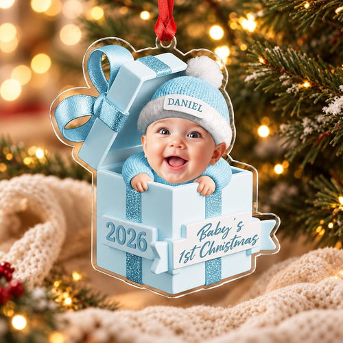 A clear acrylic Christmas ornament shaped like a light blue gift box with a baby wearing a blue knit hat and sweater inside. The ornament features the text DANIEL, 2026, and Baby's 1st Christmas, with a red ribbon for hanging.