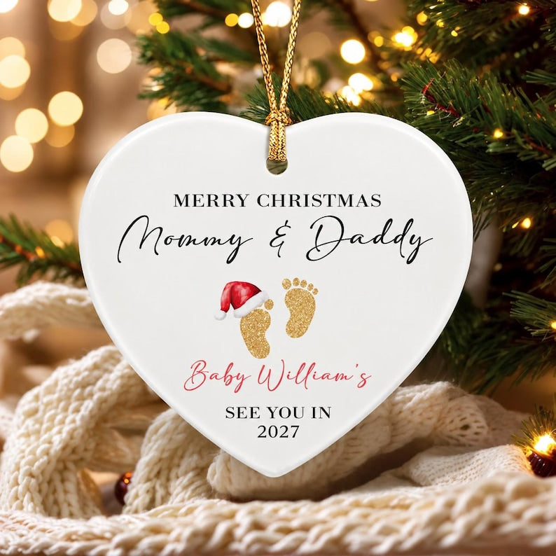 A white heart-shaped ceramic ornament with a gold hanging string. It features the black text MERRY CHRISTMAS Mommy & Daddy, gold glitter baby footprints with a red and white Santa hat, and the text Baby William's SEE YOU IN 2027.