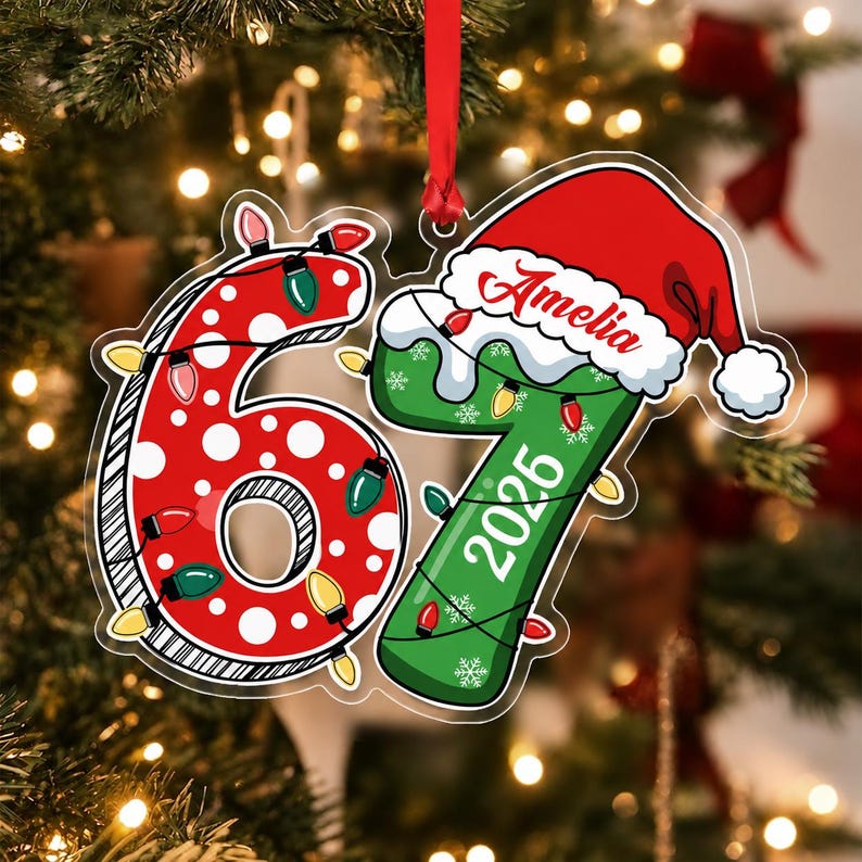 A flat, clear acrylic Christmas ornament shaped like the number 67. The 6 is red with white polka dots, and the 7 is green with white snowflakes. Both numbers are wrapped in colorful string lights.