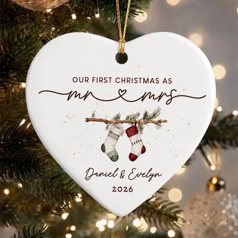 A white heart-shaped ceramic ornament featuring the text "OUR FIRST CHRISTMAS AS mr & mrs" in dark brown, with a graphic of two hanging stockings on a branch, the names "Daniel & Evelyn", and the year "2026" in brown.