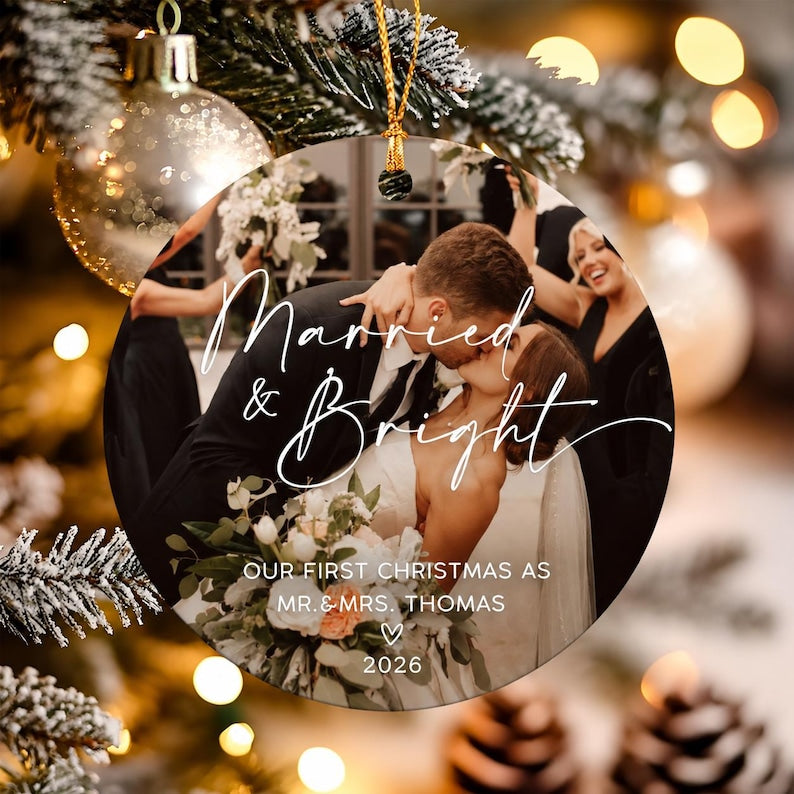 A round, flat Christmas ornament featuring a photograph of a couple kissing at their wedding. The text reads Married & Bright, OUR FIRST CHRISTMAS AS MR. & MRS. THOMAS, a heart symbol, and the year 2026.