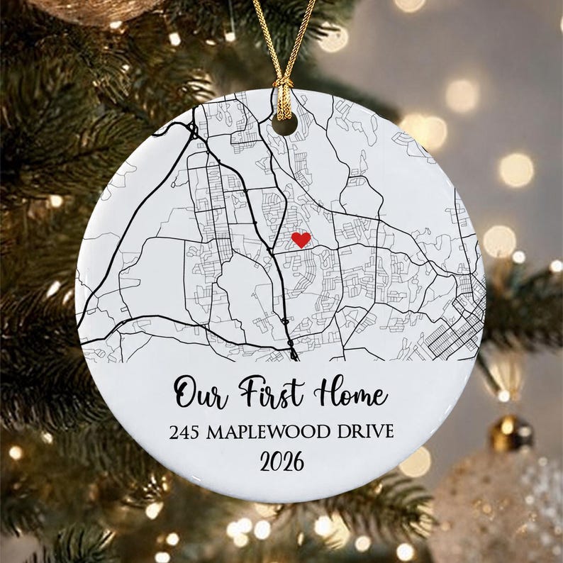 A round white ceramic ornament featuring a black line street map with a small red heart icon. The text reads Our First Home, 245 MAPLEWOOD DRIVE, and 2026. It hangs from a gold string against a blurred evergreen tree background.