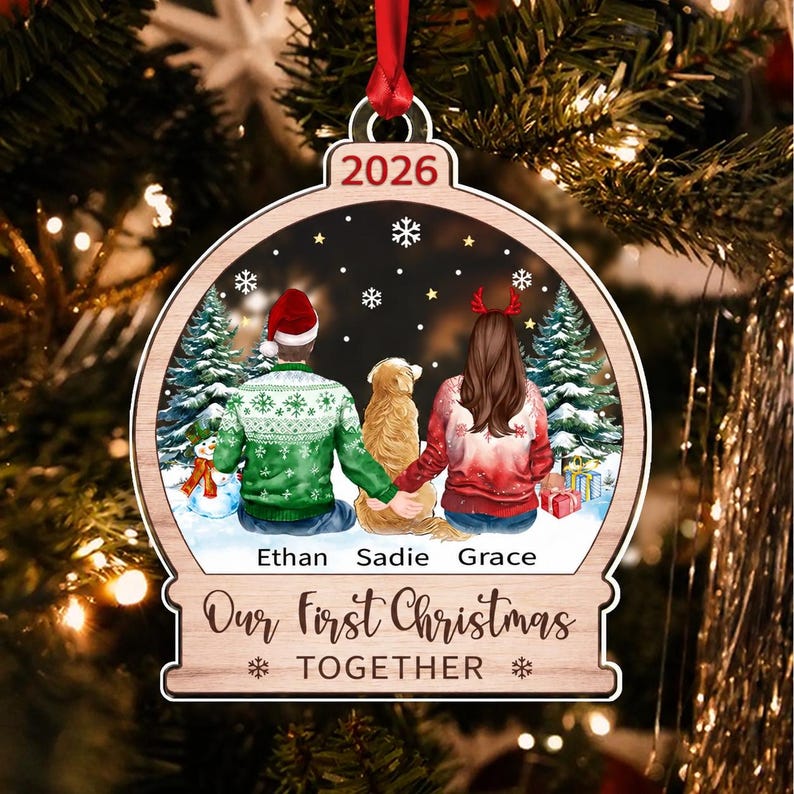 A wooden Christmas ornament shaped like a snow globe featuring a printed illustration of two people and a dog sitting in the snow. The text reads 2026, Ethan, Sadie, Grace, and Our First Christmas TOGETHER.
