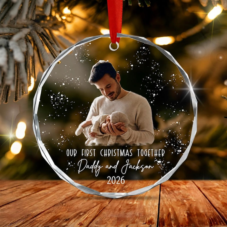 A circular, faceted glass ornament featuring a printed photograph of a person holding a baby. The text reads: OUR FIRST CHRISTMAS TOGETHER, Daddy and Jackson, 2026. A red ribbon is attached to the top for hanging.
