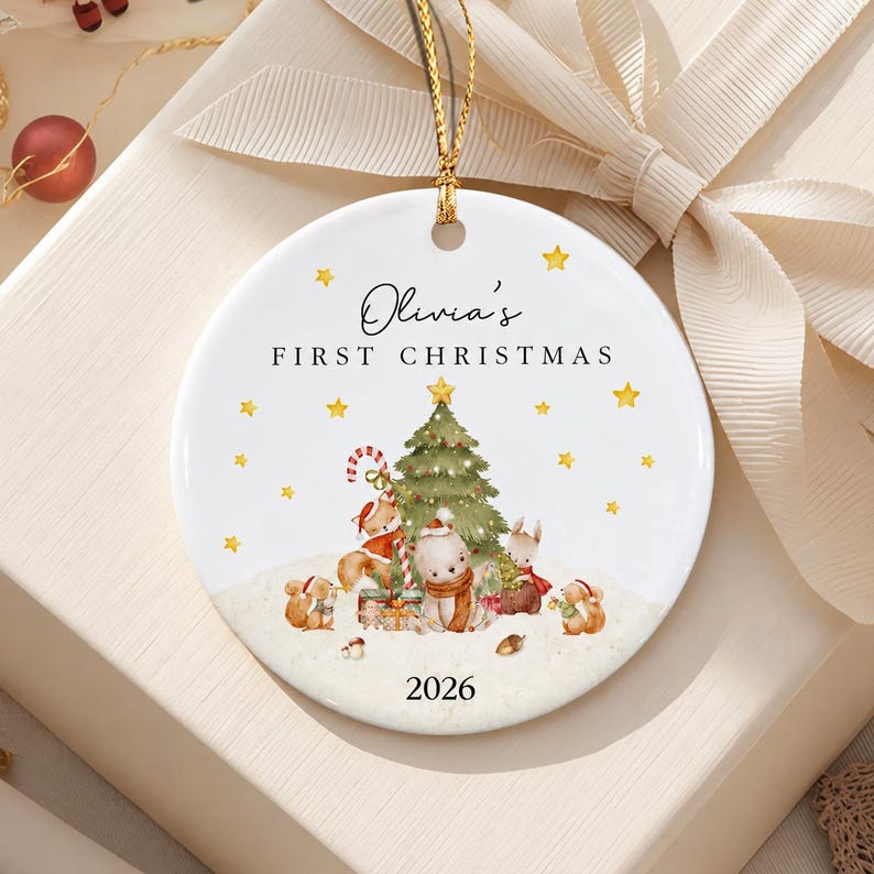 A round white ceramic ornament featuring a gold hanging string. The design includes the text Olivia's FIRST CHRISTMAS and 2026, with a central green Christmas tree, yellow stars, and illustrations of a fox, bear, and rabbit.