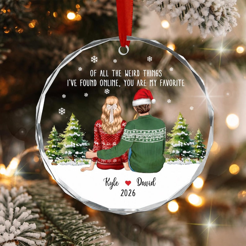 A round, faceted glass Christmas ornament featuring a couple sitting in snow with pine trees. The text reads: OF ALL THE WEIRD THINGS I'VE FOUND ONLINE, YOU ARE MY FAVORITE. Below are the names Kyle and David with 2026.