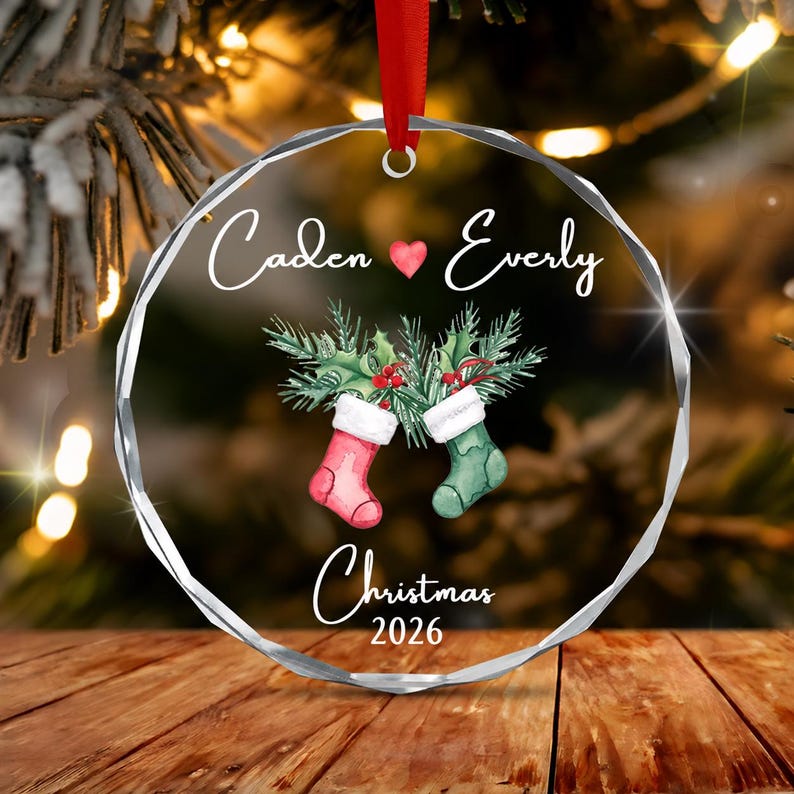 A round, faceted clear glass Christmas ornament hanging from a red ribbon. It features the text Caden, a small pink heart, and Everly, with an illustration of two stockings, one pink and one green, and the text Christmas 2026.